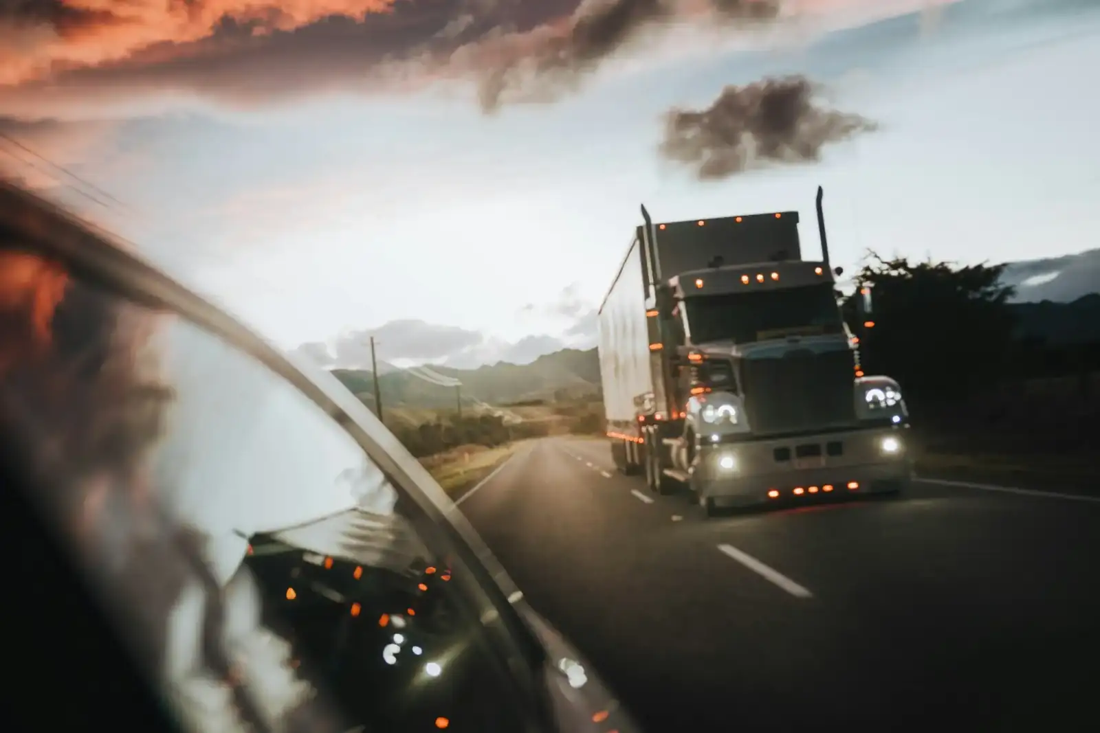 A North American semi-truck with its headlights on, approaching on an open highway under an evening sky
